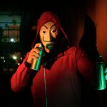 A masked person in a red hoodie holds spray paints, creating street art at night.