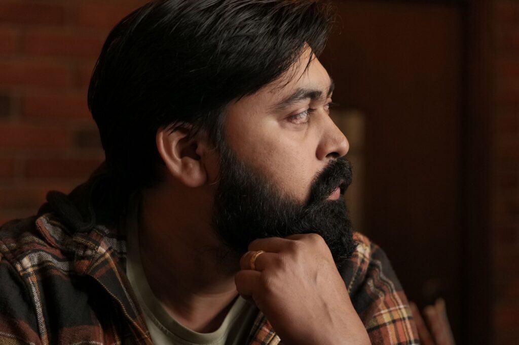 Profile portrait of a bearded South Asian man deep in thought, wearing casual plaid indoors.