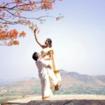 A joyful Indian couple in traditional attire celebrates love under blooming branches in a sunny landscape.