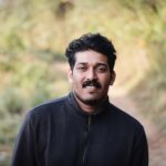 A portrait of a South Asian man in a black jacket standing outdoors on a natural pathway.