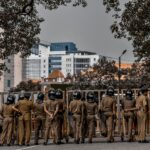 Back view of police officers in uniform standing on a city street surrounded by buildings and trees.