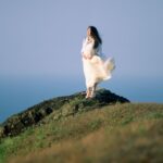 Woman in a flowing white dress standing gracefully on a hilltop, overlooking the sea.
