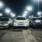 Night scene in a well-lit parking lot featuring various parked cars and street lamps.