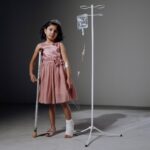 Injured girl in pink dress using crutches next to an IV stand in a studio setting.