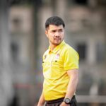 Focused soccer coach wearing yellow shirt during outdoor training session in Hanoi, Vietnam.