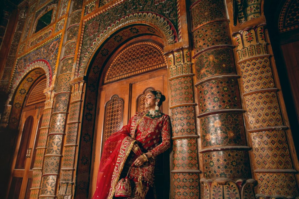 A woman in traditional red attire posing by a highly ornamented wall with intricate patterns.