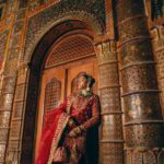 A woman in traditional red attire posing by a highly ornamented wall with intricate patterns.