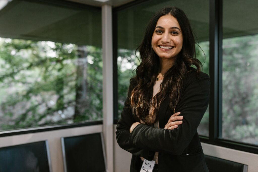 Smiling woman in business attire stands confidently in a modern office with large windows.