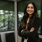 Smiling woman in business attire stands confidently in a modern office with large windows.