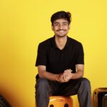 A cheerful young man in casual wear smiling against a vibrant yellow background. Indoors, positive vibe.