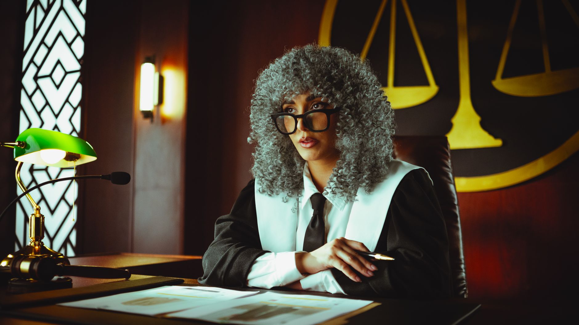 Female judge in courtroom setting, sitting at desk with justice scales in background.