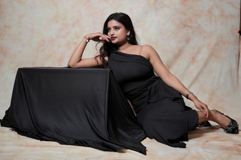 A glamorous woman in an elegant black dress poses indoors, exuding confidence and style.