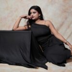 A glamorous woman in an elegant black dress poses indoors, exuding confidence and style.