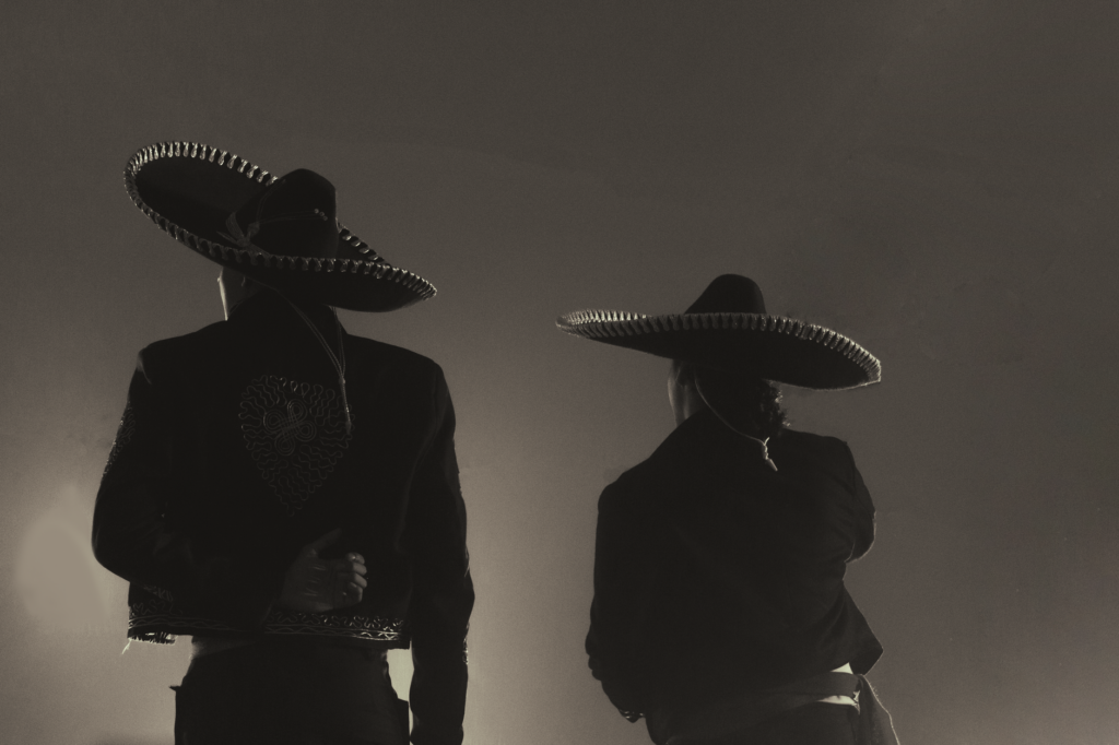 Back view of two charros in traditional costumes silhouetted in monochrome lighting.
