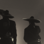Back view of two charros in traditional costumes silhouetted in monochrome lighting.