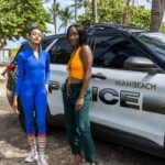Two stylish women stand beside a Miami Beach police car in a sunny outdoor setting.