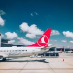 Airplanes on a tarmac at an airport with red tail logos, clear blue sky, and scattered clouds.