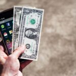 Close-up of a hand holding US dollar bills and a smartphone outdoors, showcasing financial technology.