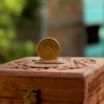 Close-up of a golden coin resting on a carved wooden piggy bank in an outdoor setting.