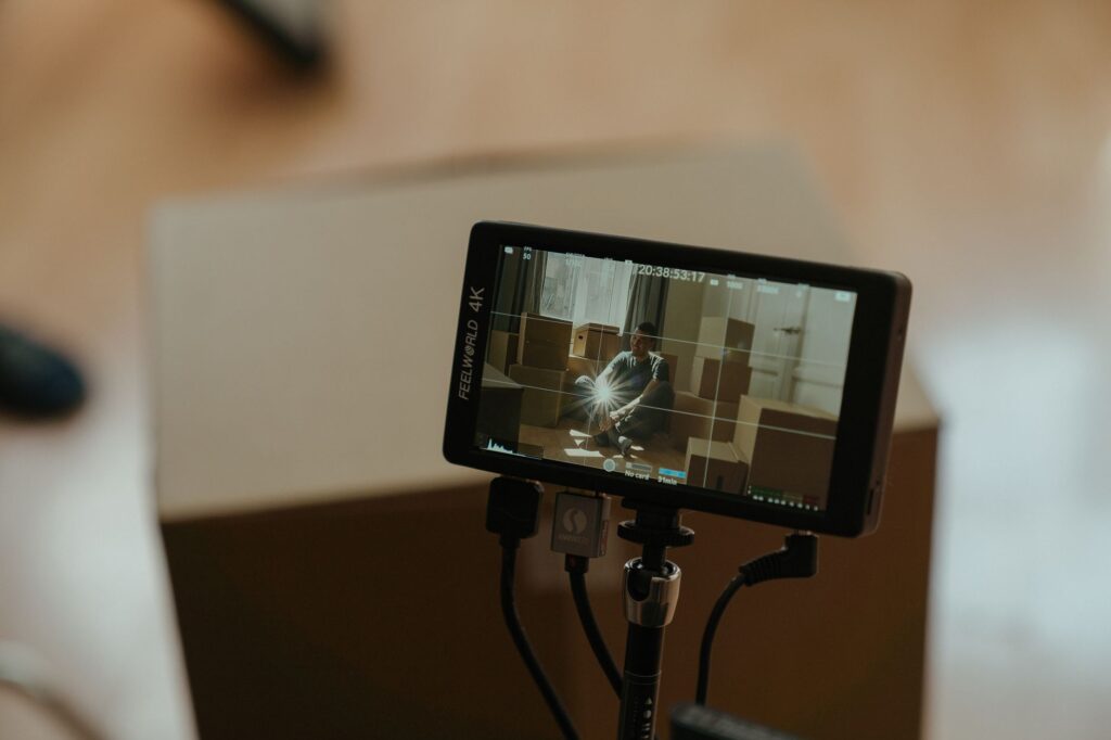 A camera monitor shows a scene with a person sitting among boxes, highlighting the filming process.