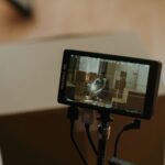 A camera monitor shows a scene with a person sitting among boxes, highlighting the filming process.