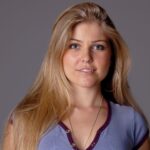 Caucasian woman with long blonde hair, smiling against a gray background.