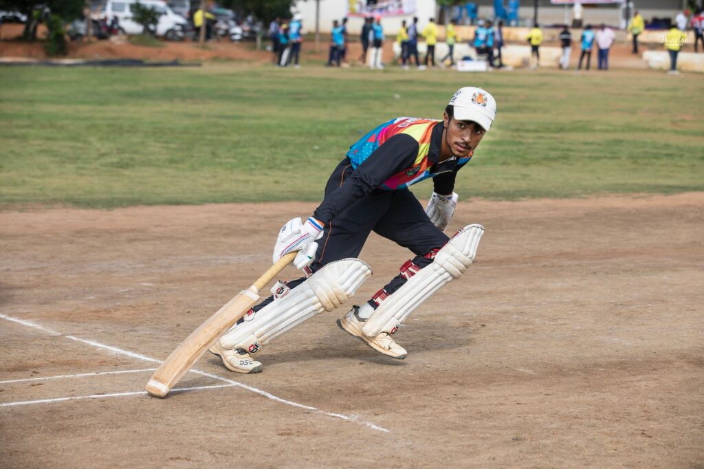 A cricket player in colorful attire bats during a daytime match on a sunny field.