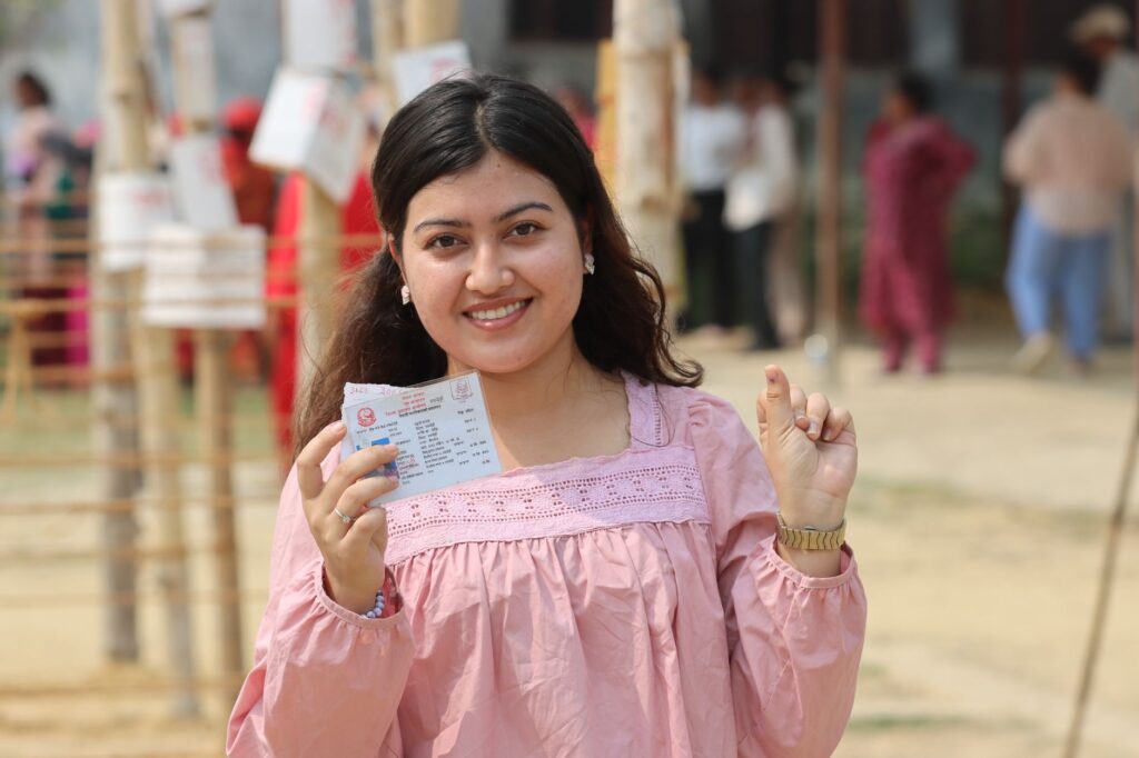 Young woman proudly displays voting ticket at outdoor polling station in Nepal.