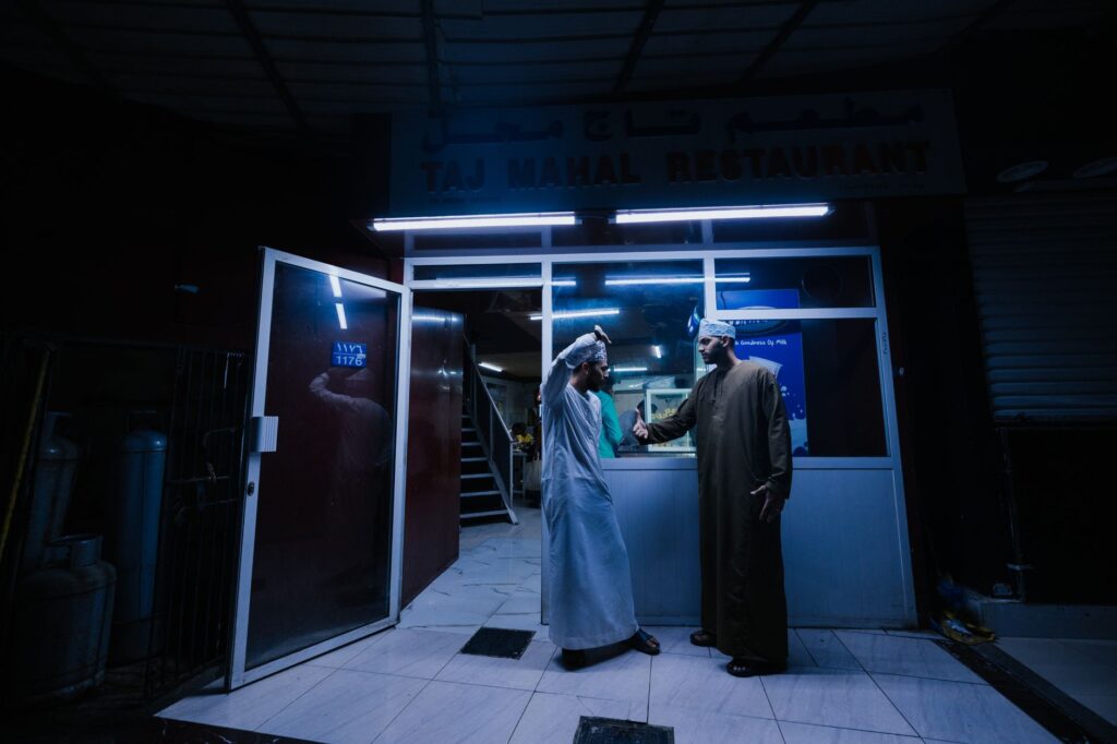 Two men engage in conversation outside the Taj Mahal Restaurant in Muscat at night.