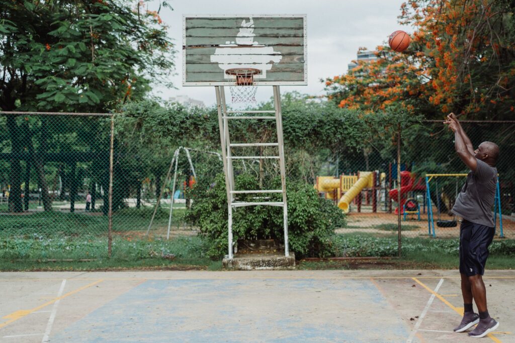 An African American man shoots a basketball in an outdoor court surrounded by lush greenery.