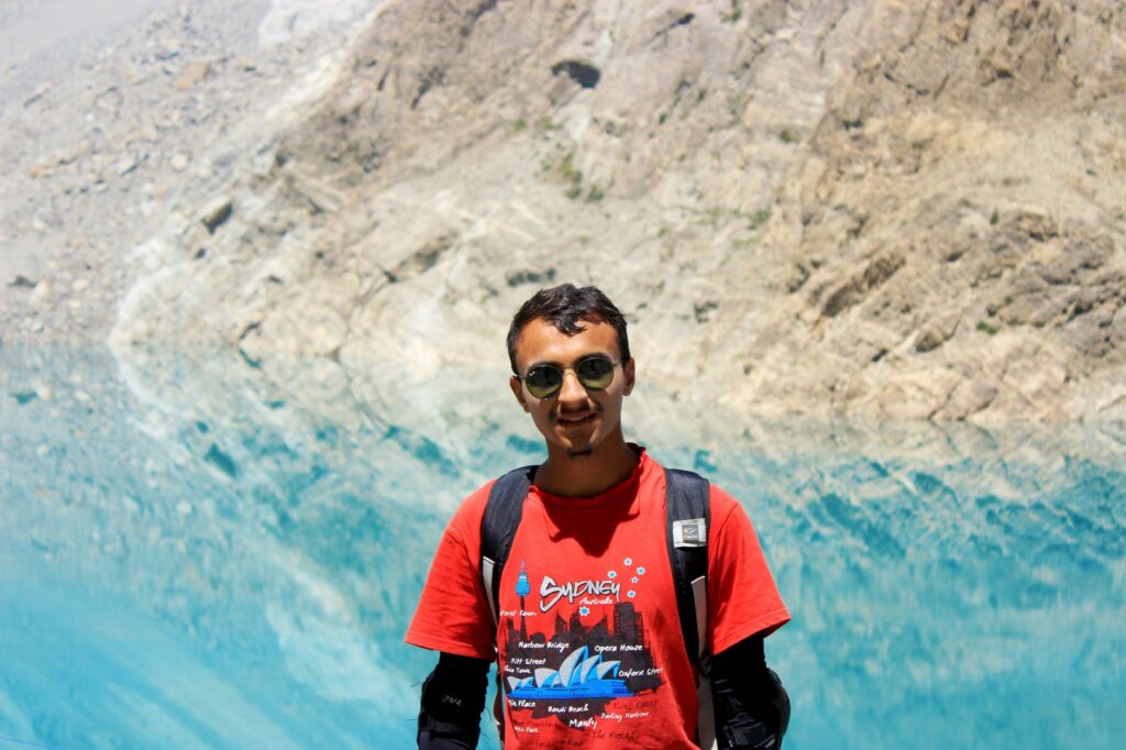A young man wearing sunglasses and a backpack enjoys the scenic view by a stunning mountain lake.