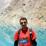 A young man wearing sunglasses and a backpack enjoys the scenic view by a stunning mountain lake.