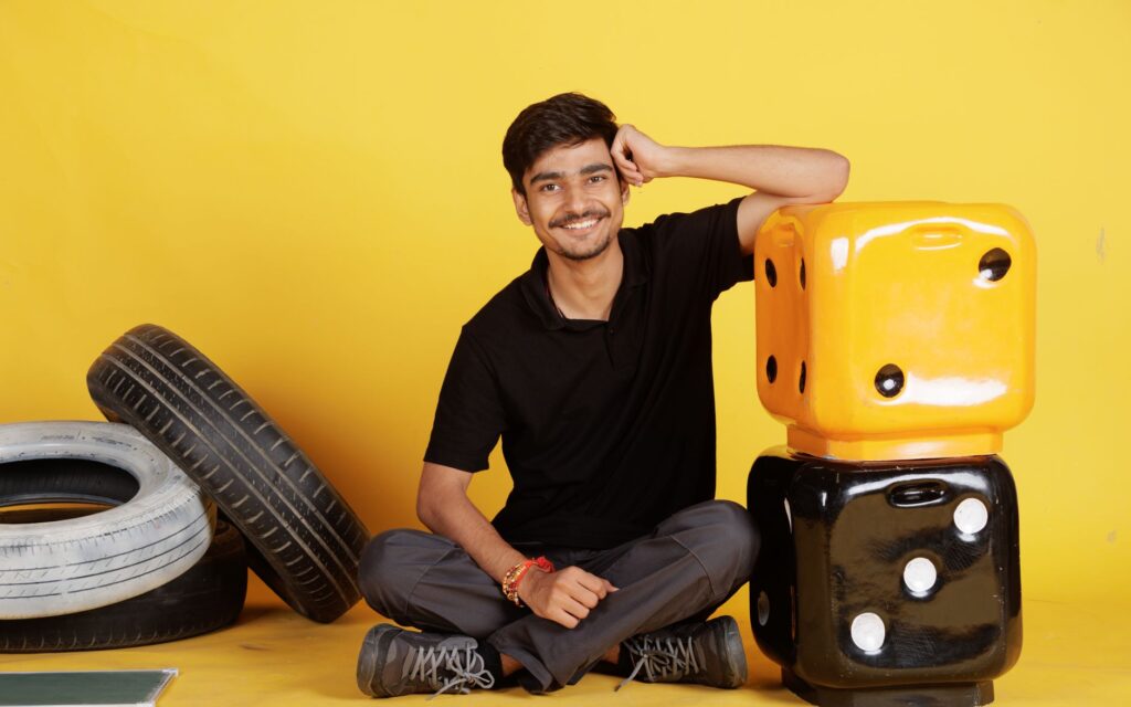 Smiling young man seated indoors with large dice and tires, exuding a positive and relaxed vibe.