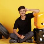 Smiling young man seated indoors with large dice and tires, exuding a positive and relaxed vibe.