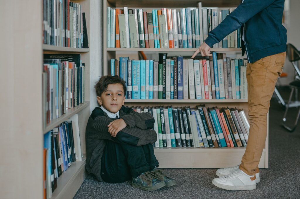 A sad boy sitting on the library floor while being bullied by a peer.