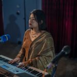 A woman playing a keyboard on stage with a microphone, wearing a saree.