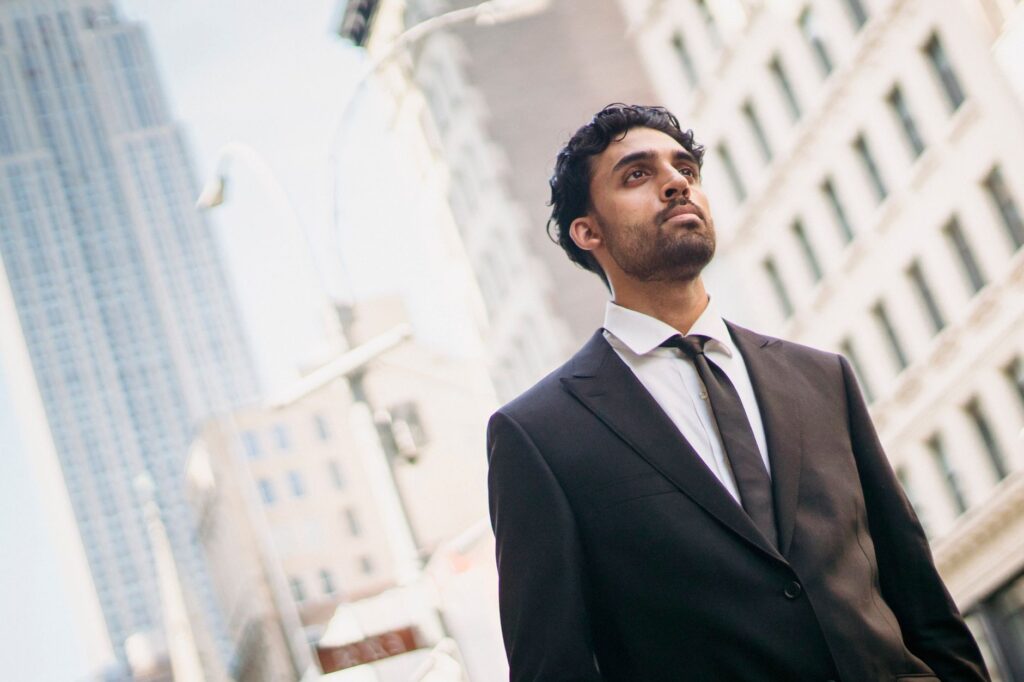 A bearded businessman in a suit stands confidently outdoors against a cityscape.