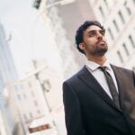A bearded businessman in a suit stands confidently outdoors against a cityscape.