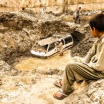 A child watches as a van is trapped in flood debris in Madyan, Pakistan, highlighting the aftermath of natural disaster.