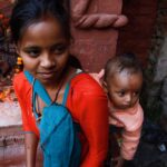 A South Asian woman carrying her baby in traditional attire at an ornate temple setting.