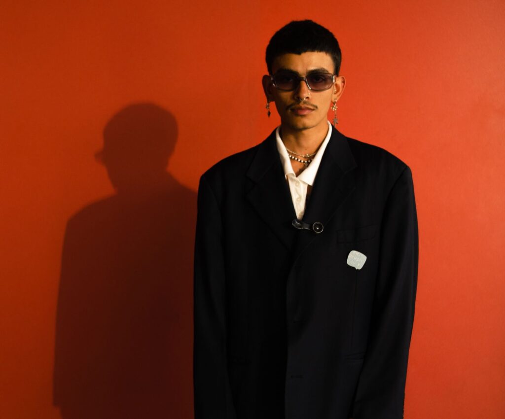 Fashion portrait of a trendy man posing with sunglasses and jewelry against a vibrant red background.
