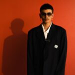 Fashion portrait of a trendy man posing with sunglasses and jewelry against a vibrant red background.
