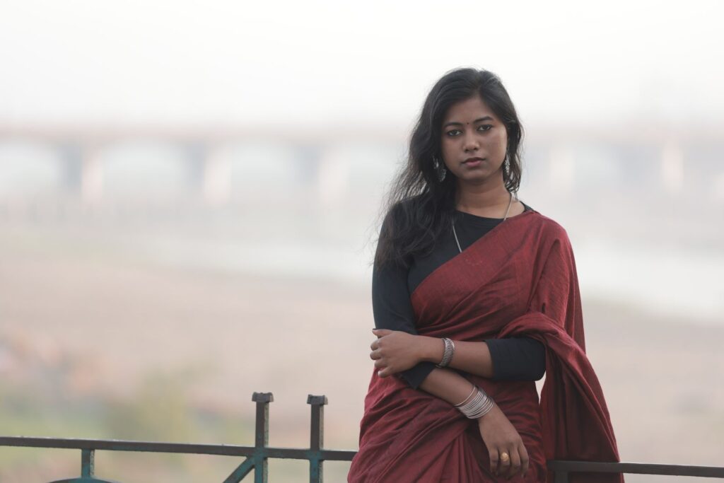 An elegant outdoor portrait of a woman in a red saree with a blurred bridge in the background.