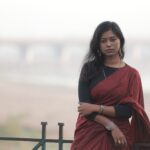 An elegant outdoor portrait of a woman in a red saree with a blurred bridge in the background.