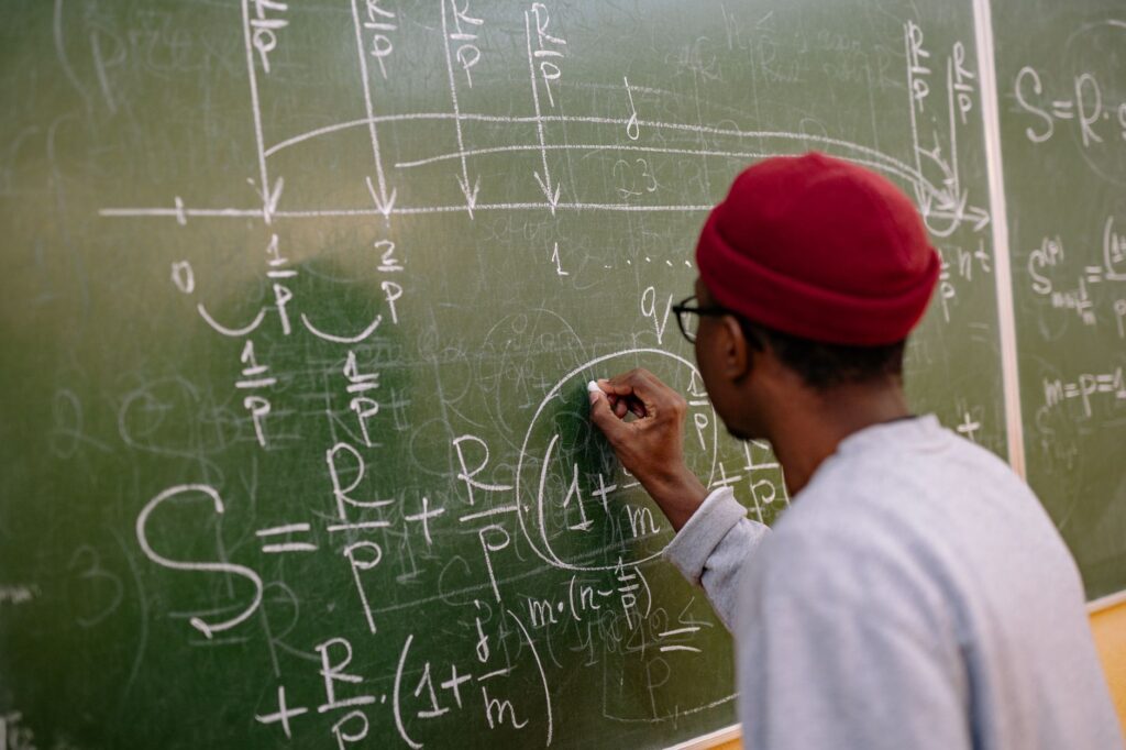 Student writing complex mathematical equations on a chalkboard in a classroom setting.
