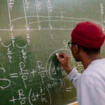 Student writing complex mathematical equations on a chalkboard in a classroom setting.