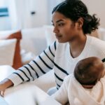 Ethnic baby on laps hugging female in casual outfit with netbook at table while sitting in chair at light apartment near sofa