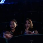 Two women sitting in a dark cinema, enjoying popcorn and watching a movie.