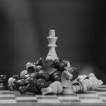 Black and white image of a chess king standing victorious amid fallen pieces on a board.