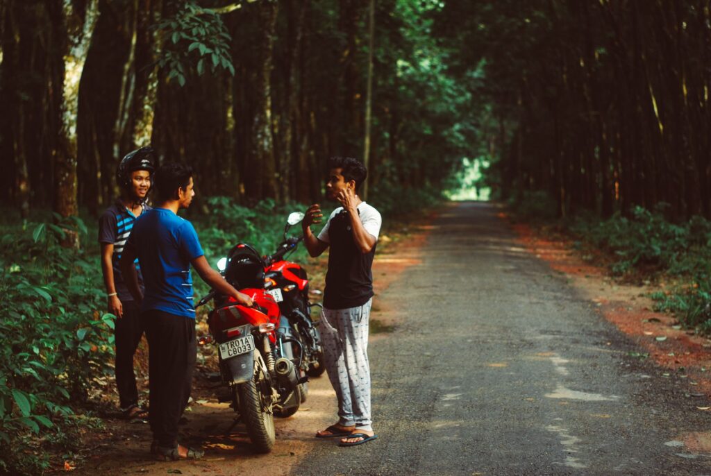 A casual group of friends chatting beside motorbikes on a tranquil forest road in Mainama, India.